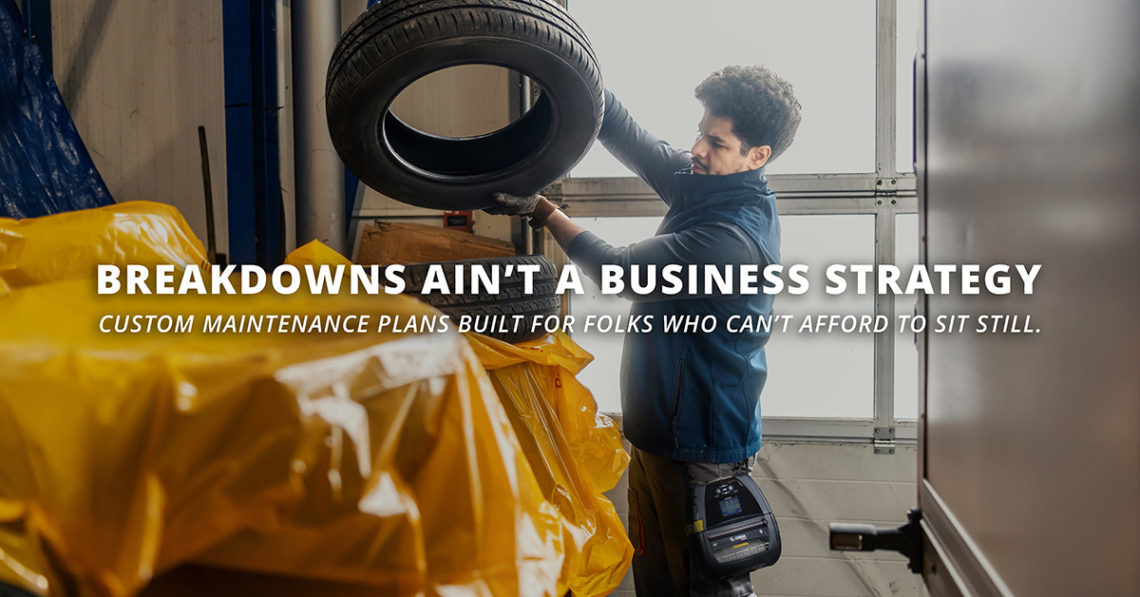 Warehouse worker lifting a tire with a rugged Zebra device on his hip, standing beside wrapped equipment. Bold white text reads “Breakdowns Ain’t a Business Strategy” with a subheading promoting custom maintenance plans. Image promotes tailored support and custom maintenance agreements to enhance operational efficiency.