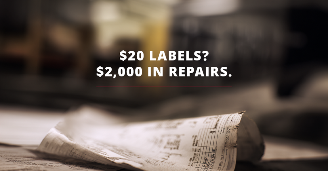 A broken, damaged label laying on a tabletop in a warehouse. Overlaid copy reads "$20 Labels? $2,000 in repairs>"