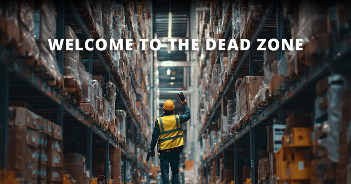 Man walking through warehouse in hi-vis vest and hard hat, holding a mobile device above his head to find signal. Text overlay reads "welcome to the dead zone"